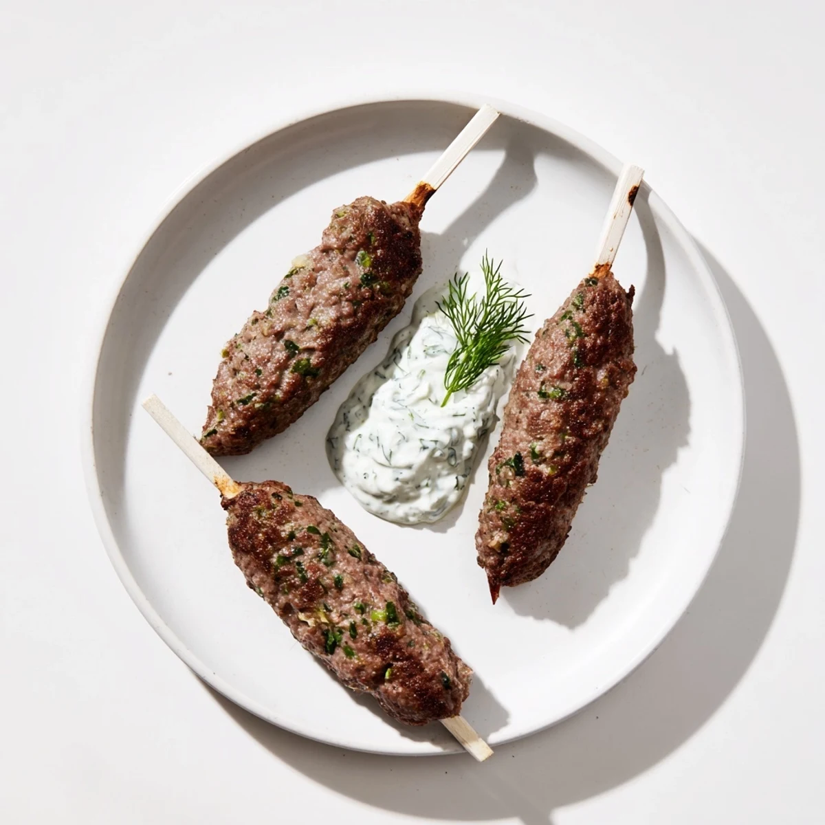 Juicy grilled beef kofta kebabs topped with creamy cucumber yogurt sauce and fresh herbs on a plate.