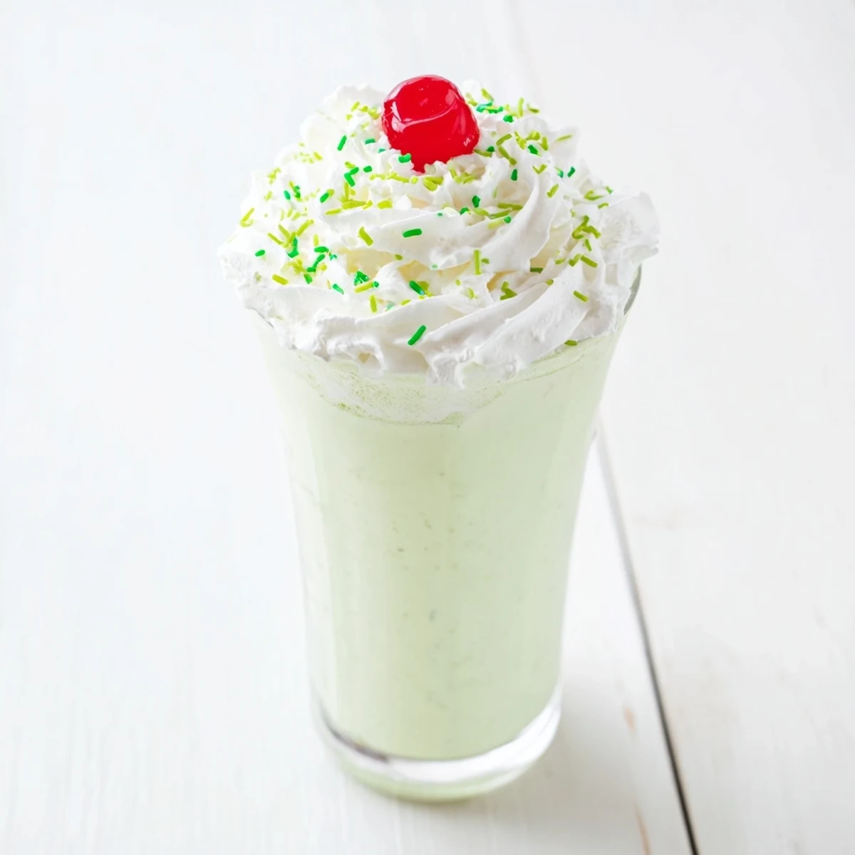 Close-up of a frothy, mint-green Homemade Copycat Shamrock Shake in a glass, garnished with whipped cream and green sprinkles for a sweet treat.