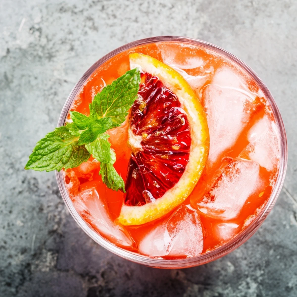 Vibrant red-hued Blood Orange Lemonade in a clear glass with ice cubes, garnished with blood orange wheels and a sprig of mint on a wooden table.
