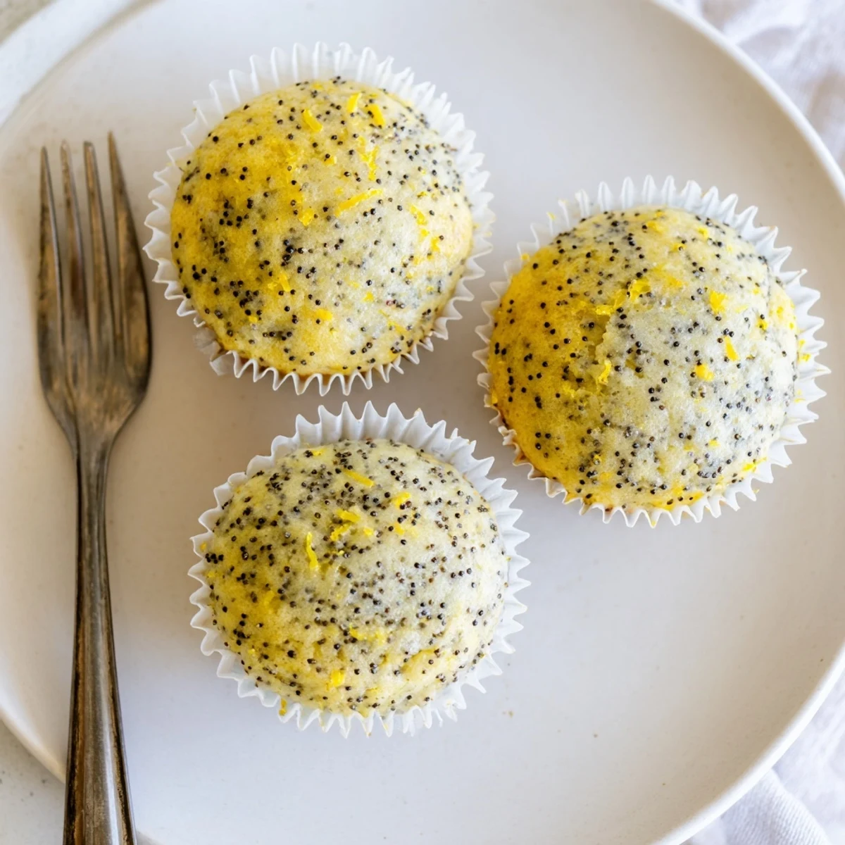 Moist lemon poppy seed muffins studded with tiny black seeds beside a bowl of bright zest.